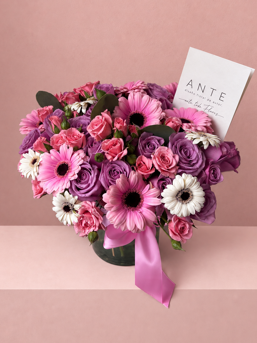 Arreglo floral con rosas moradas, gerberas rosas y blancas, y rosas spray en florero de vidrio bajo con moño rosa, entrega a domicilio CDMX